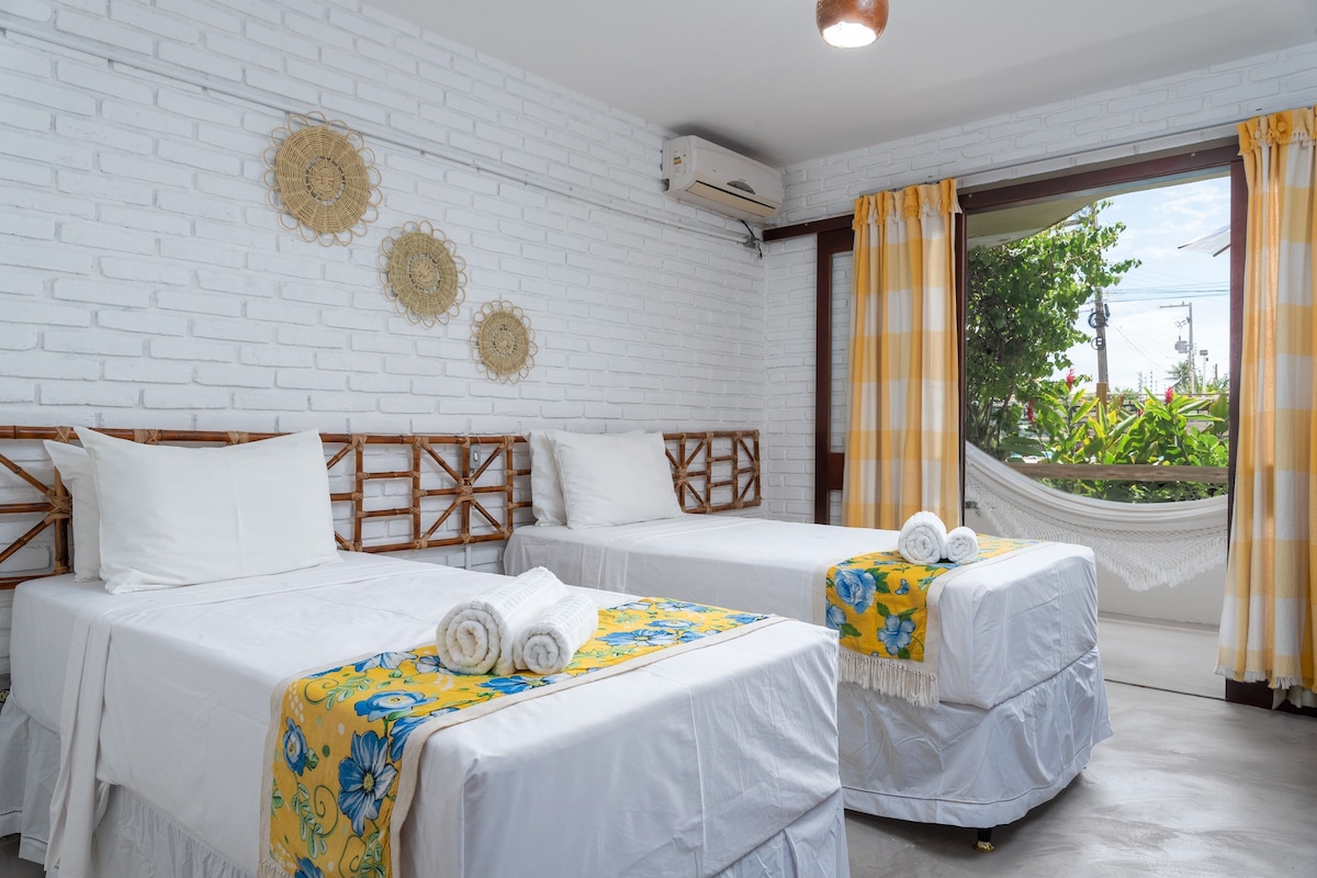 Une chambre d’hôtel avec deux lits, du linge de lit blanc, un hamac et une vue sur la verdure par une fenêtre ornée de rideaux jaunes.