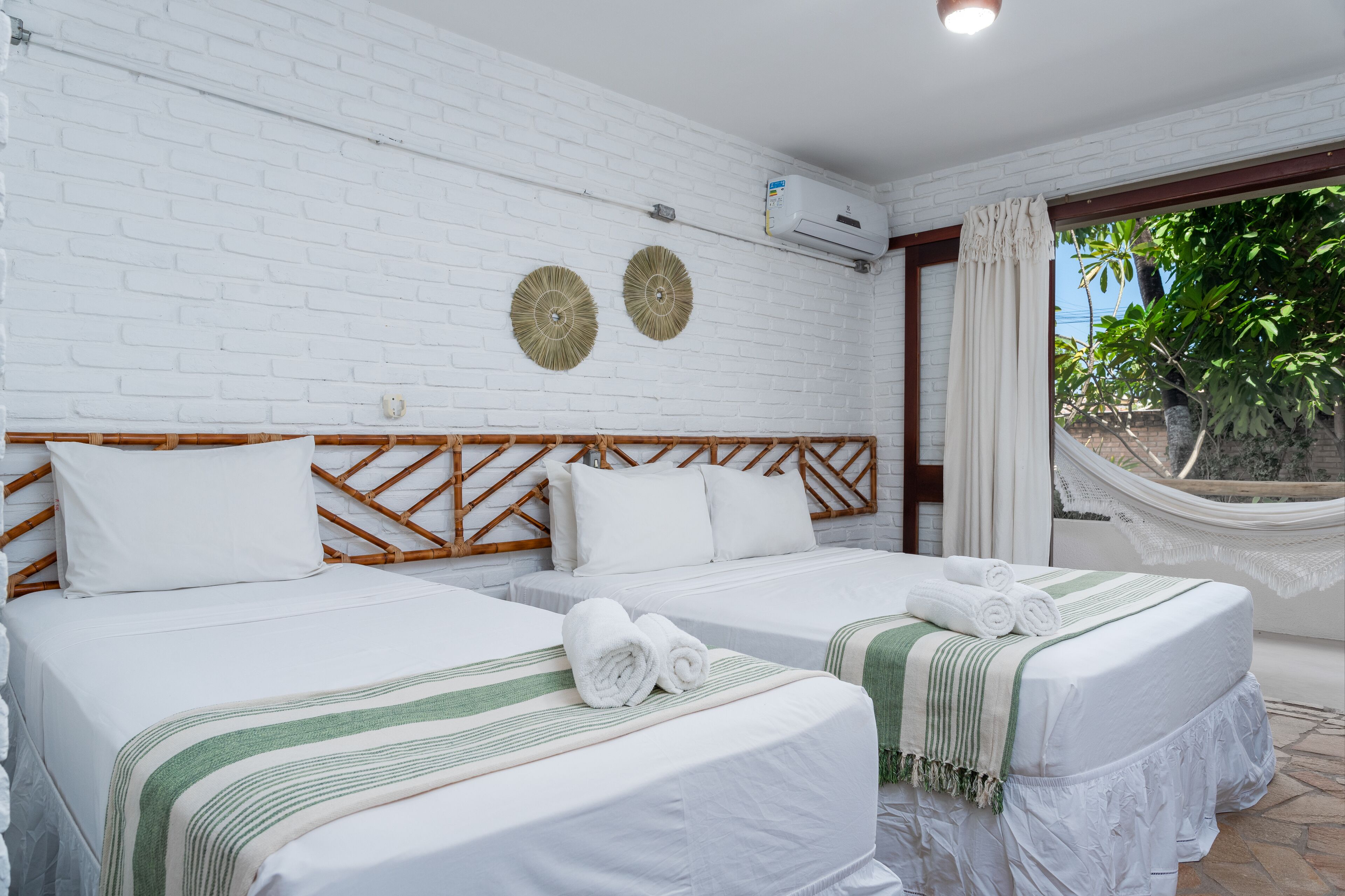 Une chambre d’hôtel avec deux lits, du linge de lit blanc, un mur en briques blanches, deux éléments de décoration murale et un hamac près de la fenêtre.
