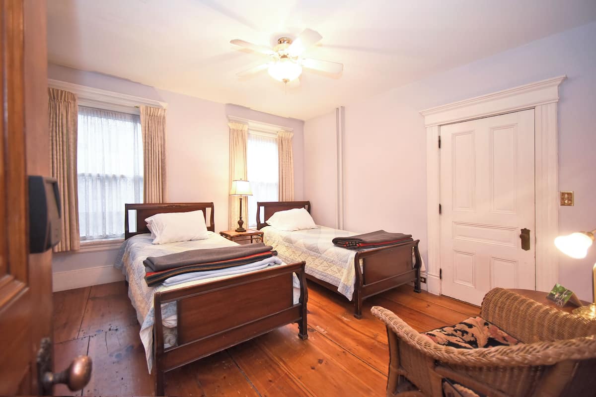 A bedroom with two wooden beds, a ceiling fan, a door, and a wicker chair.
