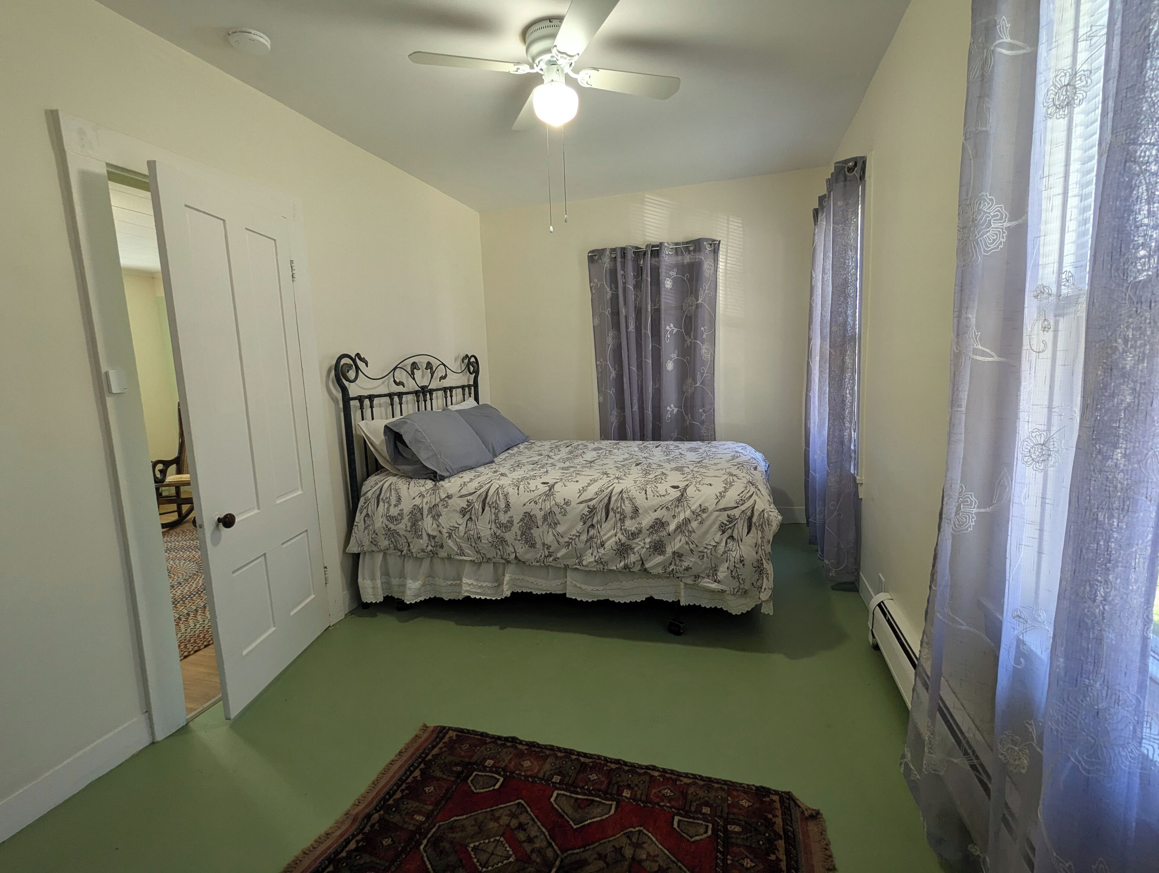 Ein Schlafzimmer mit einem Bett, einem Deckenventilator, einer Tür und einem Vorhangfenster.