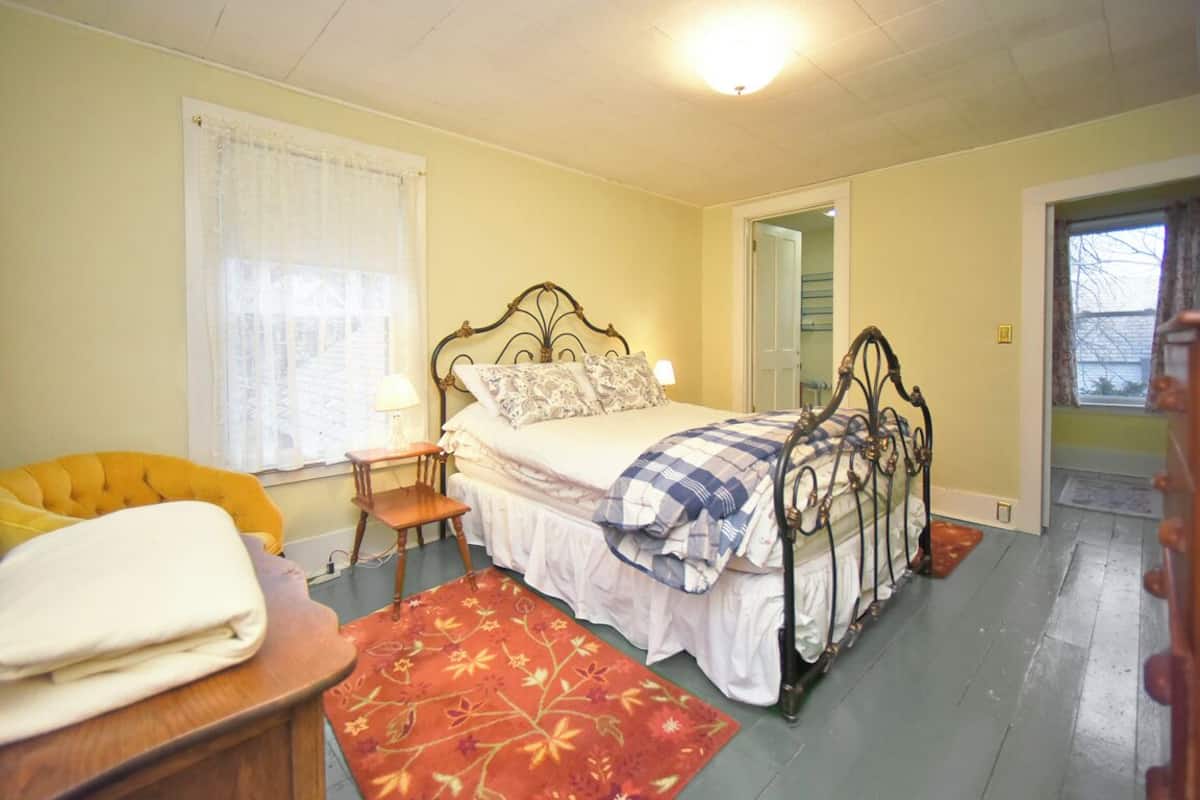 A bedroom with a bed, a chair, a nightstand, a window with curtains, and a door leading to another room.