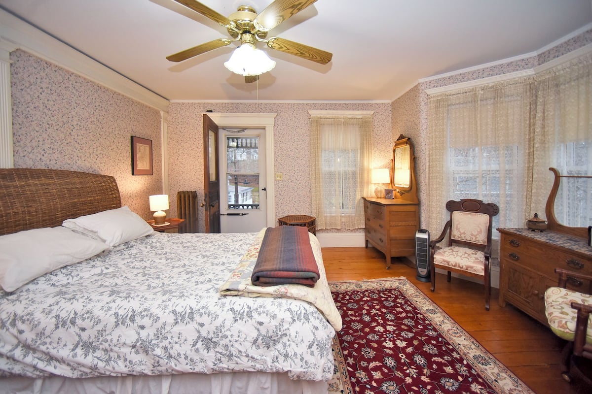 A bedroom with a large bed, a wooden dresser, a chair, a mirror, and a ceiling fan.