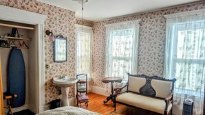 A bedroom with floral wallpaper, a bed, a dresser, a vanity, a chair, and a window with curtains. - Blooming Inn (Shelburne Falls)