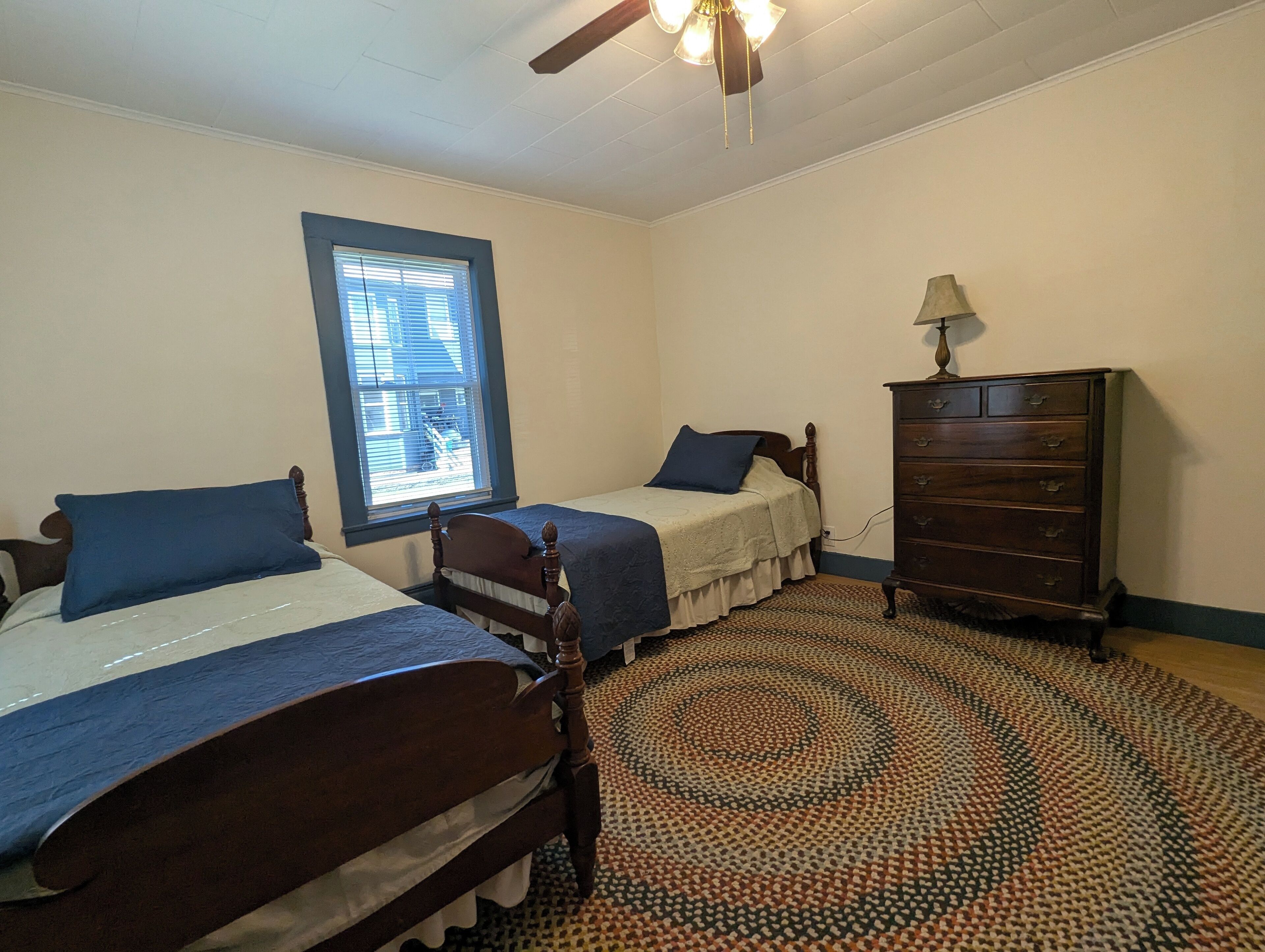 Une chambre à coucher avec deux lits en bois, une commode, un tapis à motifs et un ventilateur de plafond.