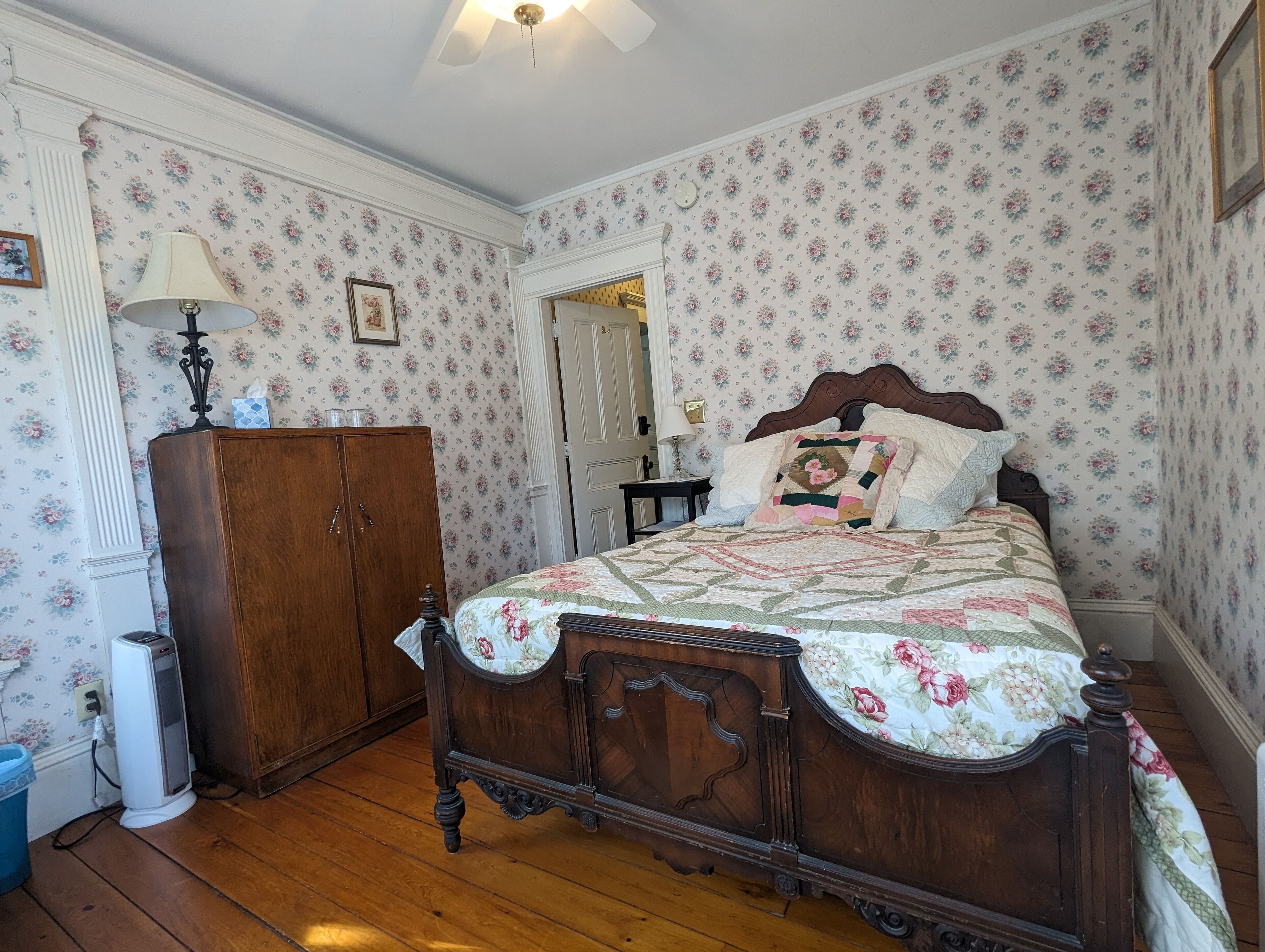 Un dormitorio con cama de madera, papel tapiz floral, un armario de madera y una mesita de noche con lámpara.