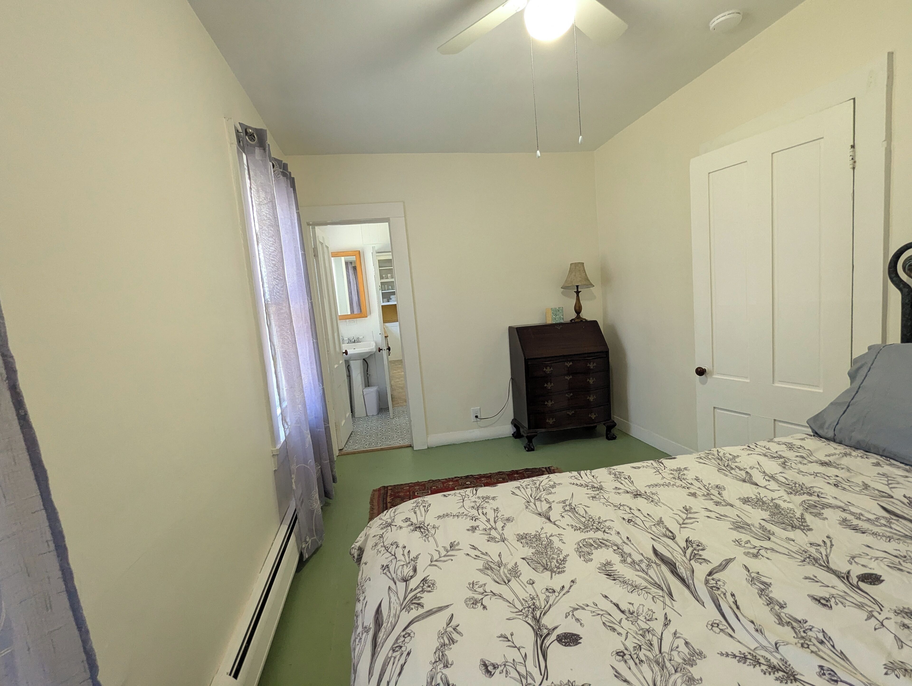 A bedroom with a bed, a dresser, a nightstand, a ceiling fan, and a door leading to a bathroom.