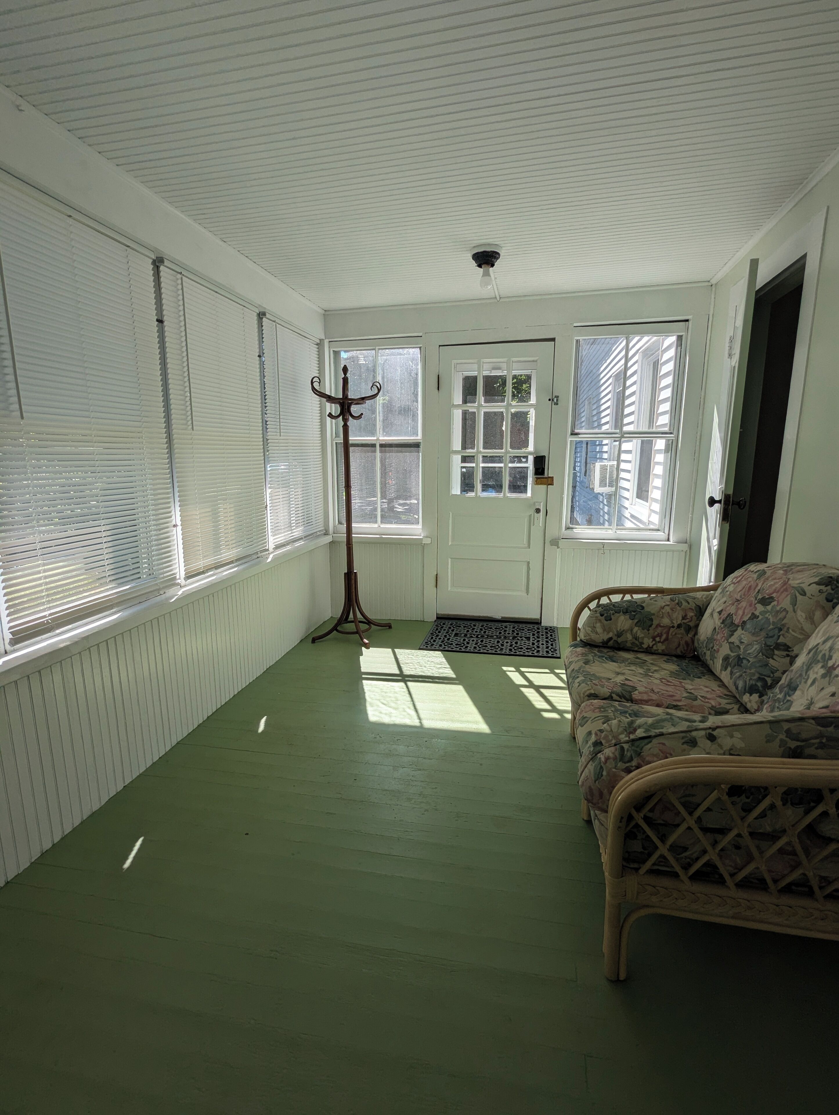 Una habitación con piso verde, un sofá de mimbre, un perchero y una puerta con ventanal.