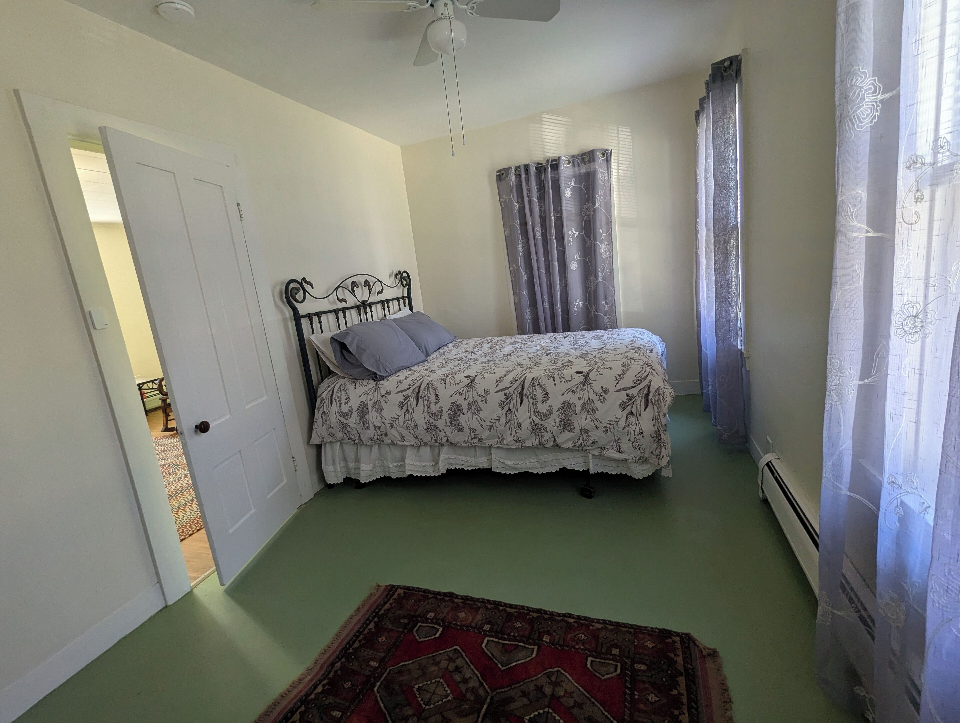 Un dormitorio con cama, ventilador de techo, ventana con cortinas y puerta que da a otra habitación.