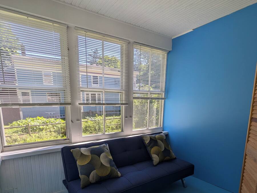 Ein Zimmer mit einem dunkelblauen Sofa, zwei gemusterten Kissen und Blick durch große Fenster mit Jalousien.