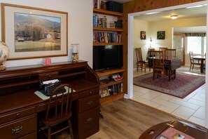 A living room with a wooden desk, bookshelves, a television, and a dining area with chairs and a table.