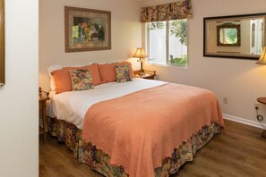 A bedroom with a large bed, floral-patterned bedding, a nightstand with a lamp, a window with a view of greenery, and a mirror on the wall.