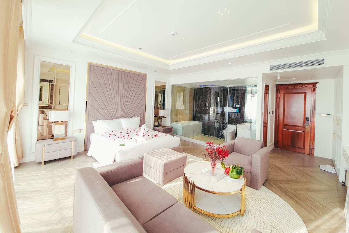 A modern hotel room with a large bed, a sitting area with a sofa and a coffee table, a bathroom with a glass-enclosed shower, and a mirror reflecting the room.