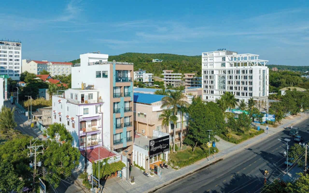 A multi-story hotel with balconies, a street view, and a hilly background.