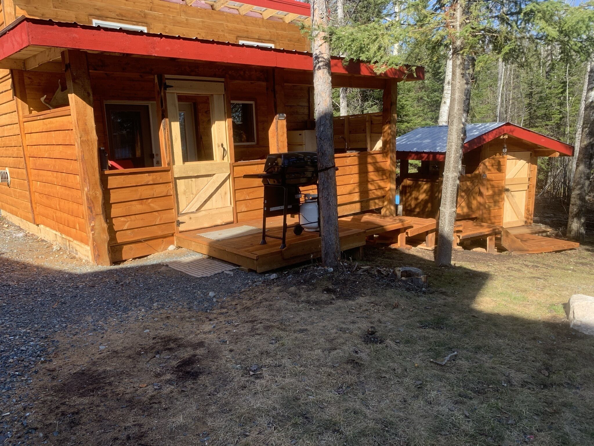 Een houten hut met een veranda, een barbecue en een kleine schuur.
