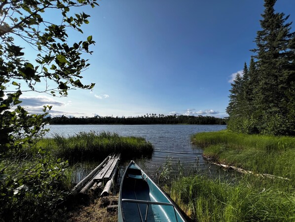 Ein ruhiger See mit einem angelegten Kanu, umgeben von dichtem Wald und blauem Himmel.