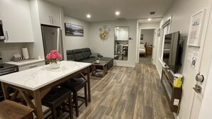 Une cuisine moderne avec un plan de travail en marbre, un coin repas avec des chaises en bois, un salon avec un canapé et une chambre à coucher visible à travers une porte ouverte.