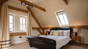 Een slaapkamer met een schuine kap, een groot raam met gordijnen, een bed met wit beddengoed, een houten nachtkastje met een lamp, en zichtbare houten balken.