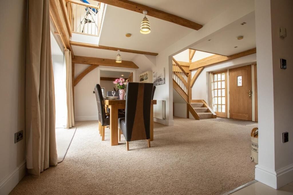A spacious room with a high ceiling, wooden beams, a dining area with a table and chairs, and a staircase leading to another level.