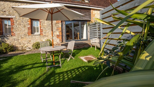 Een gebouw van steen met een houten terras, een tafel met stoelen en een grote parasol.
