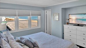 A bedroom with a bed, a dresser, a television, and a window with a beach view. - Sunrise & Sunset Oceanfront Condo (Ocean CIty)