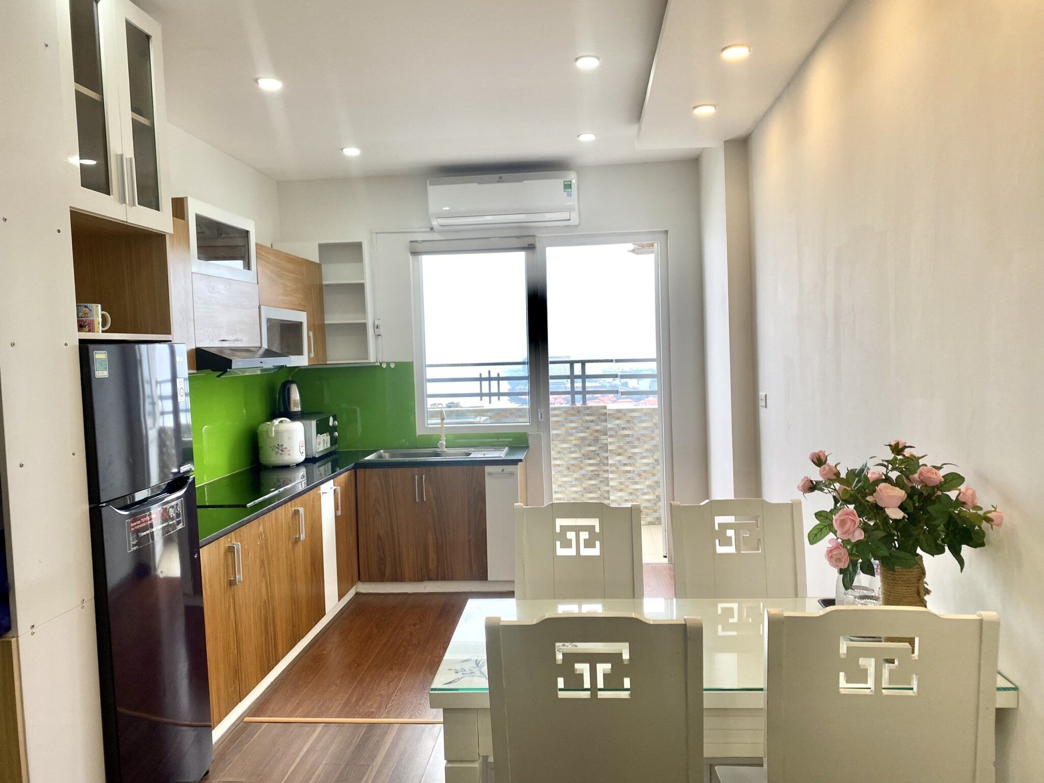 A modern kitchen with a refrigerator, microwave, and green countertop. There is a dining area with a table and chairs, and a balcony with a view.
