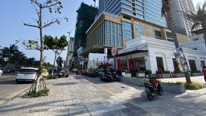 A hotel with a modern facade, a covered outdoor seating area, and a parking lot with several cars and motorcycles. - Muong Thanh Apartment Luxury 03 bedroom (Da Nang)