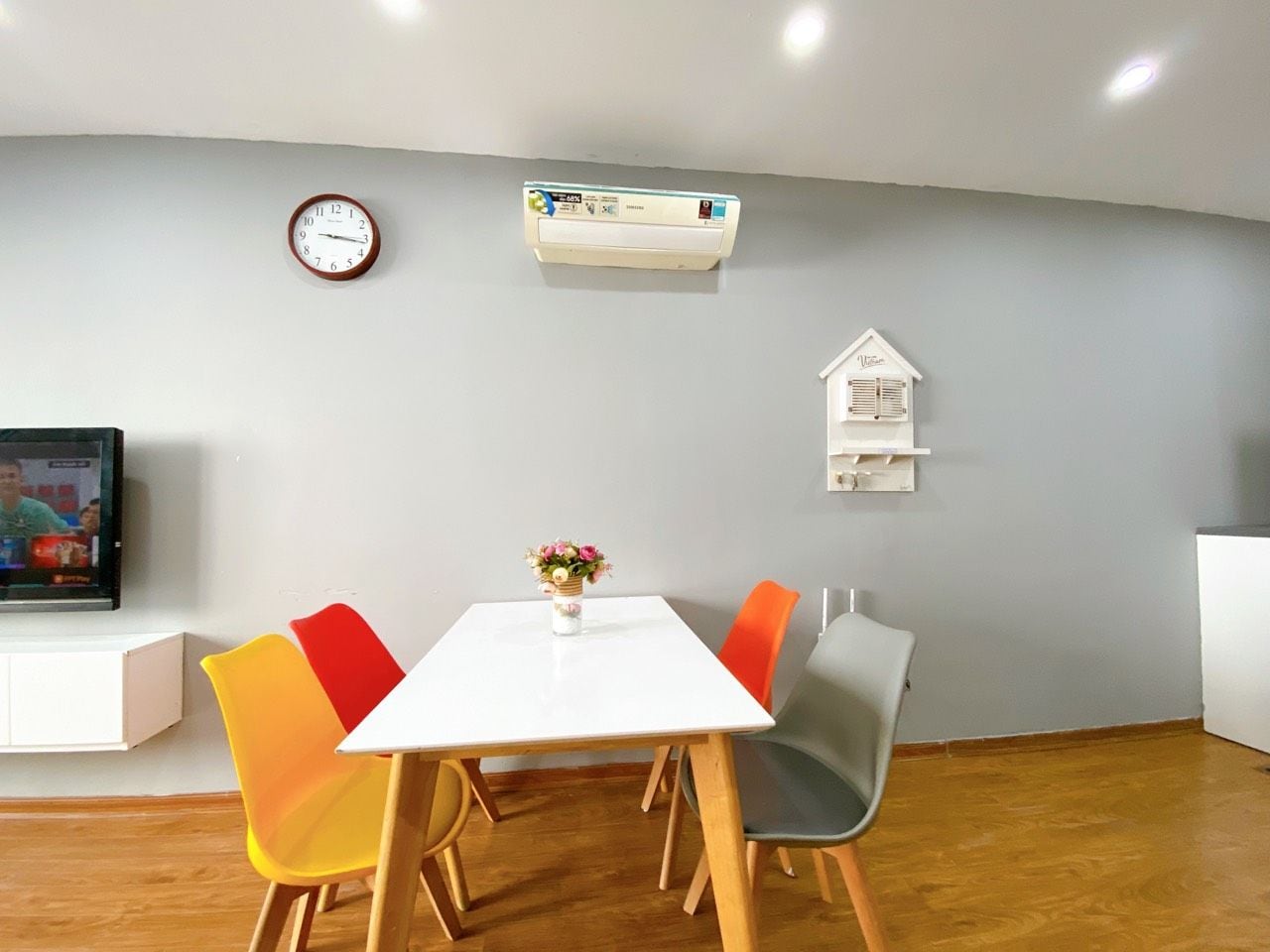 Un comedor con una mesa blanca, sillas coloridas, un reloj, un aire acondicionado y una pequeña estantería.
