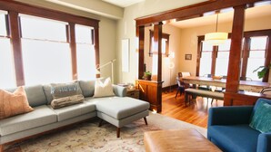 A living room with a sectional sofa, a blue armchair, a wooden dining table, and a large window with natural light. - 1912 Historic Ballard Craftsman
5 Bed • 2 Bath • Yard • Parking (Seattle)