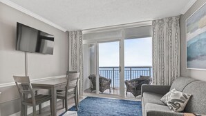 A modern living room with a flat-screen TV, a dining table with chairs, a sofa with cushions, and a large sliding glass door leading to a balcony with a view of the sea. - Amazing OCEAN FRONT Views!  Newly Renovated!   Walking Distance to All  Myrtle.  (Myrtle Beach)