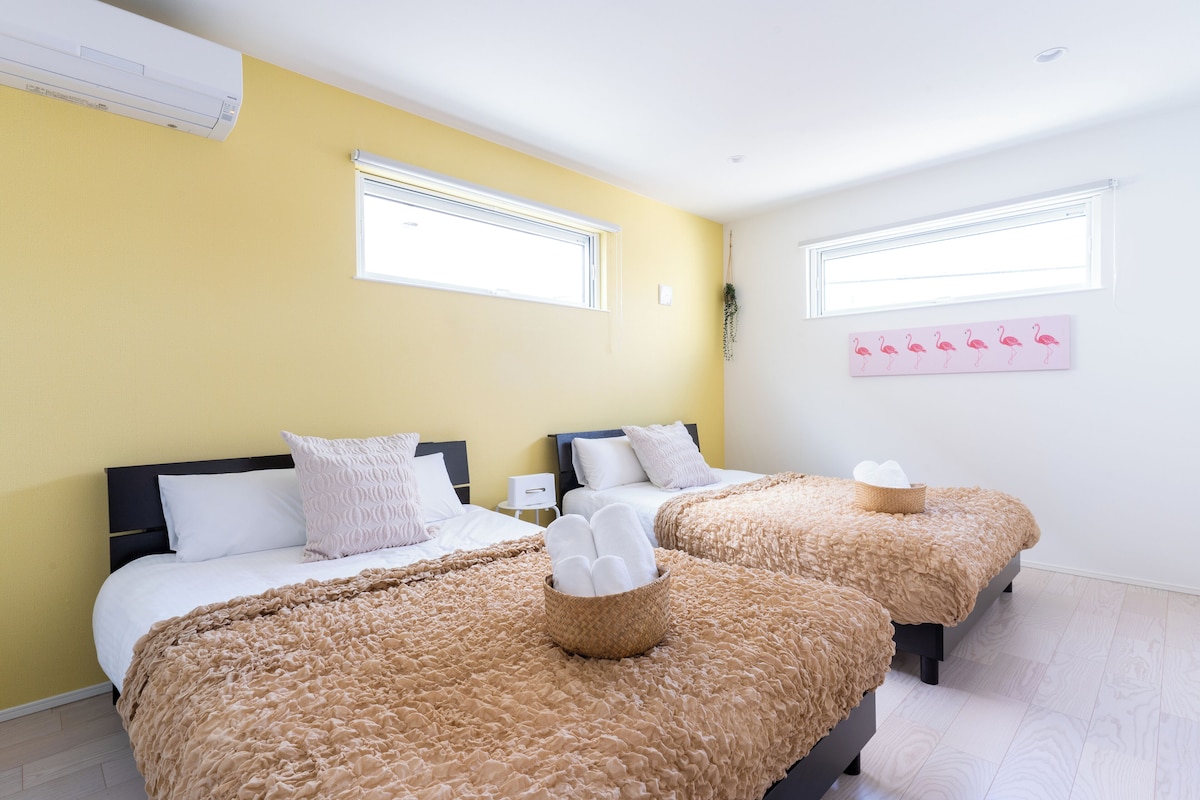 A hotel room with two beds, each with a fluffy beige blanket and a basket of white towels. There are two windows with white blinds, and a wall-mounted artwork of flamingos.