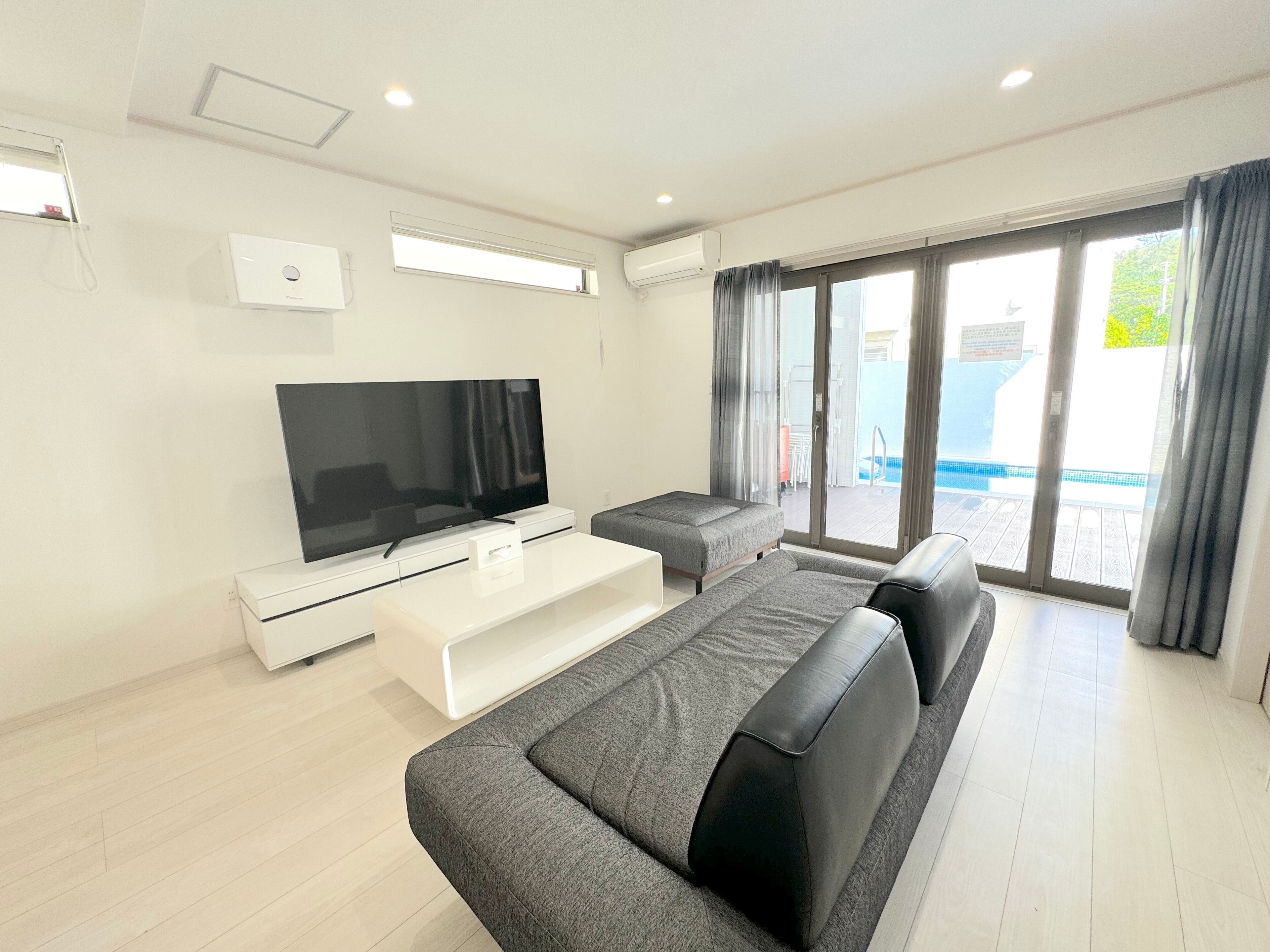 A modern living room with a flat-screen TV, a white entertainment unit, a grey sofa, and a balcony with a pool view.