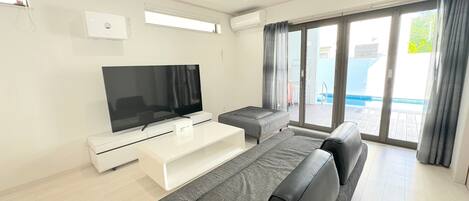 A modern living room with a flat-screen TV, a white entertainment unit, a grey sofa, and a balcony with a pool view.