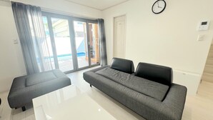 A modern living room with a grey sofa, a white coffee table, and a large sliding glass door leading to an outdoor area. - Grandioso Okinawa Pool Villa Onna 7 (Onna)