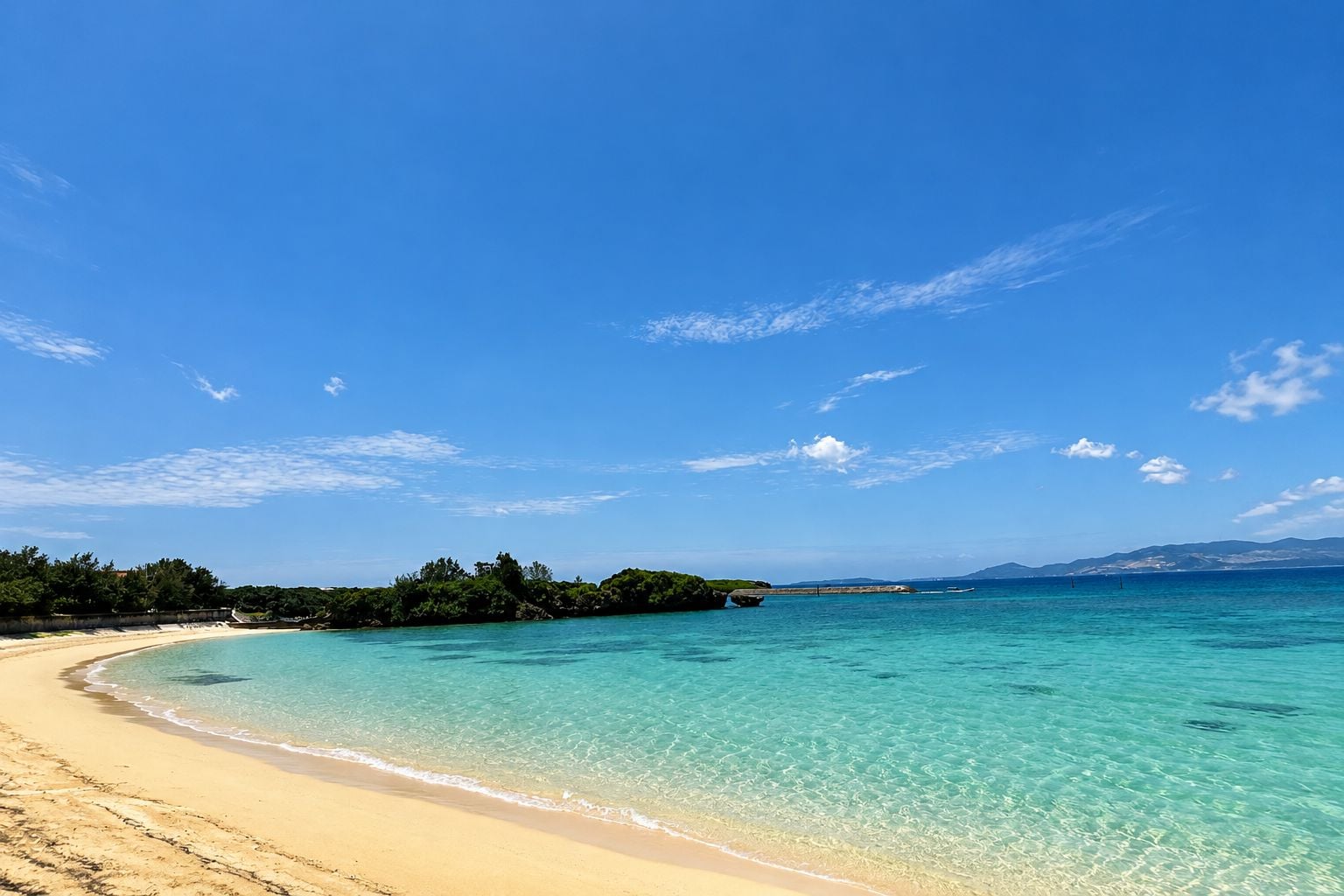 A beach with clear turquoise water, a sandy shore, and a forested island in the distance.