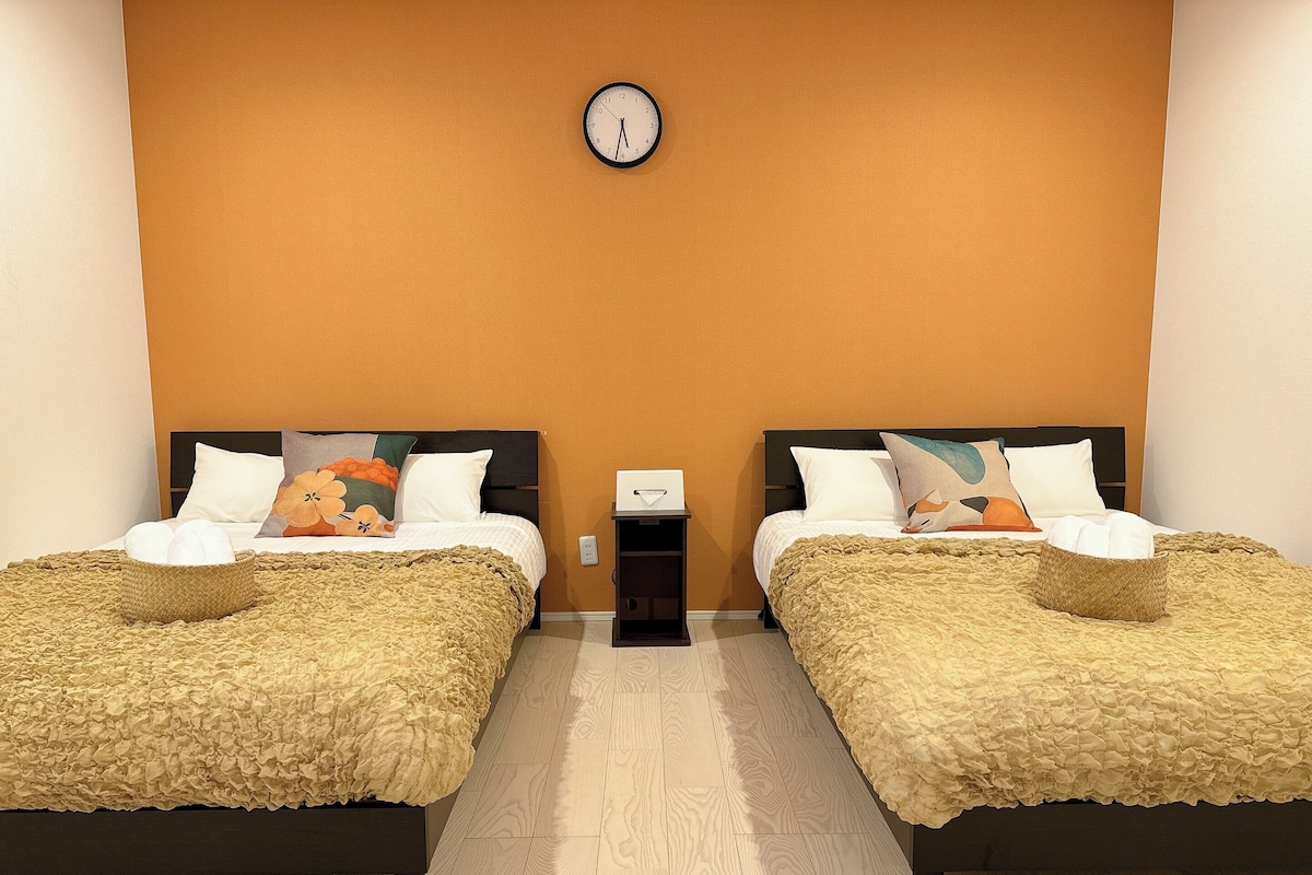 A hotel room with two beds, each with a ruffled beige blanket and pillows, a wooden headboard, and a wall clock.