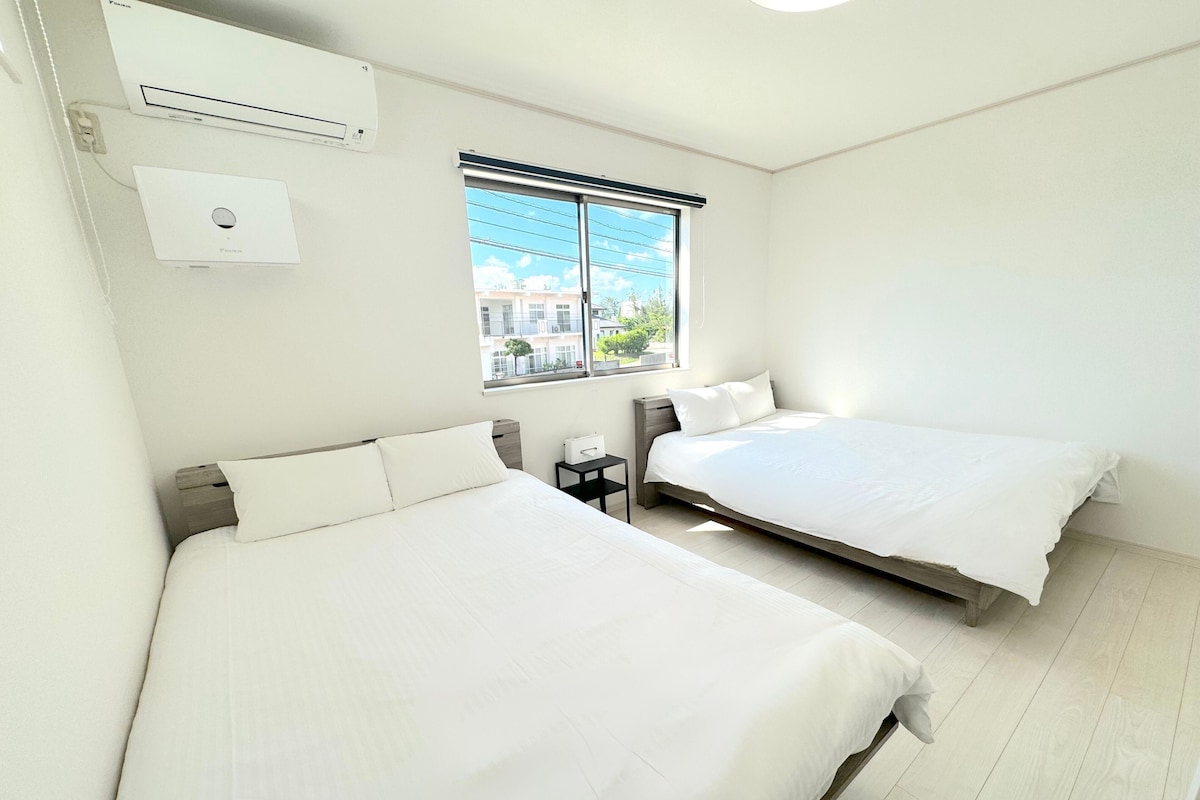 A room with two beds, a window with blinds, an air conditioning unit, and a small table.