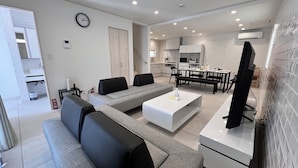 A modern living room with a sofa, coffee table, and TV. There is a kitchen area with a dining table and chairs. - Grandioso Okinawa Pool Villa Onna 7 (Onna)