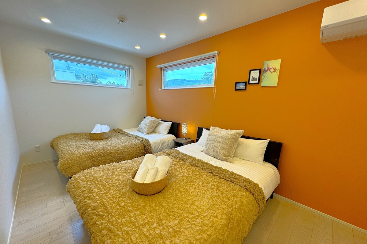A hotel room with two beds, each with a ruffled yellow bedspread, white pillows, and a small bedside table with a lamp. There are two windows with blinds, and the walls are painted orange.