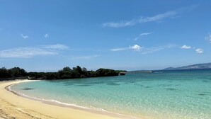 A beach with clear turquoise water, a sandy shore, and a distant view of land with vegetation and a boat. - Grandioso Okinawa Pool Villa Onna 7 (Onna)