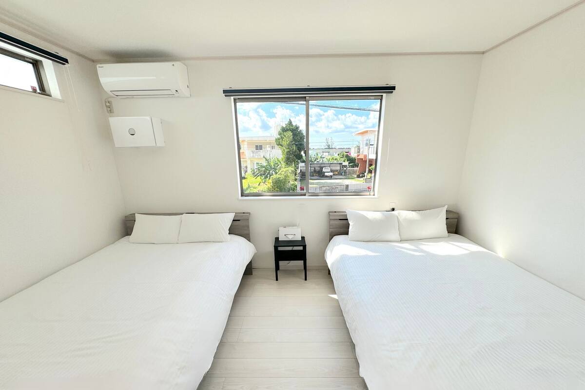 Hotellihuone, jossa on kaksi sänkyä, ikkuna, josta on näkymä rakennuksiin ja viheralueille, sekä ilmastointilaite.