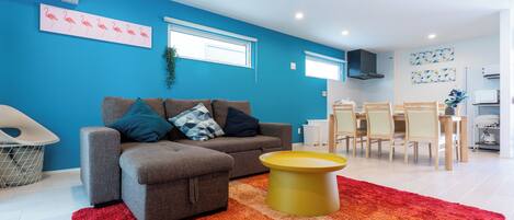 A modern living room with a grey sofa, a yellow coffee table, and a red rug. There is a dining area with a white table and chairs.