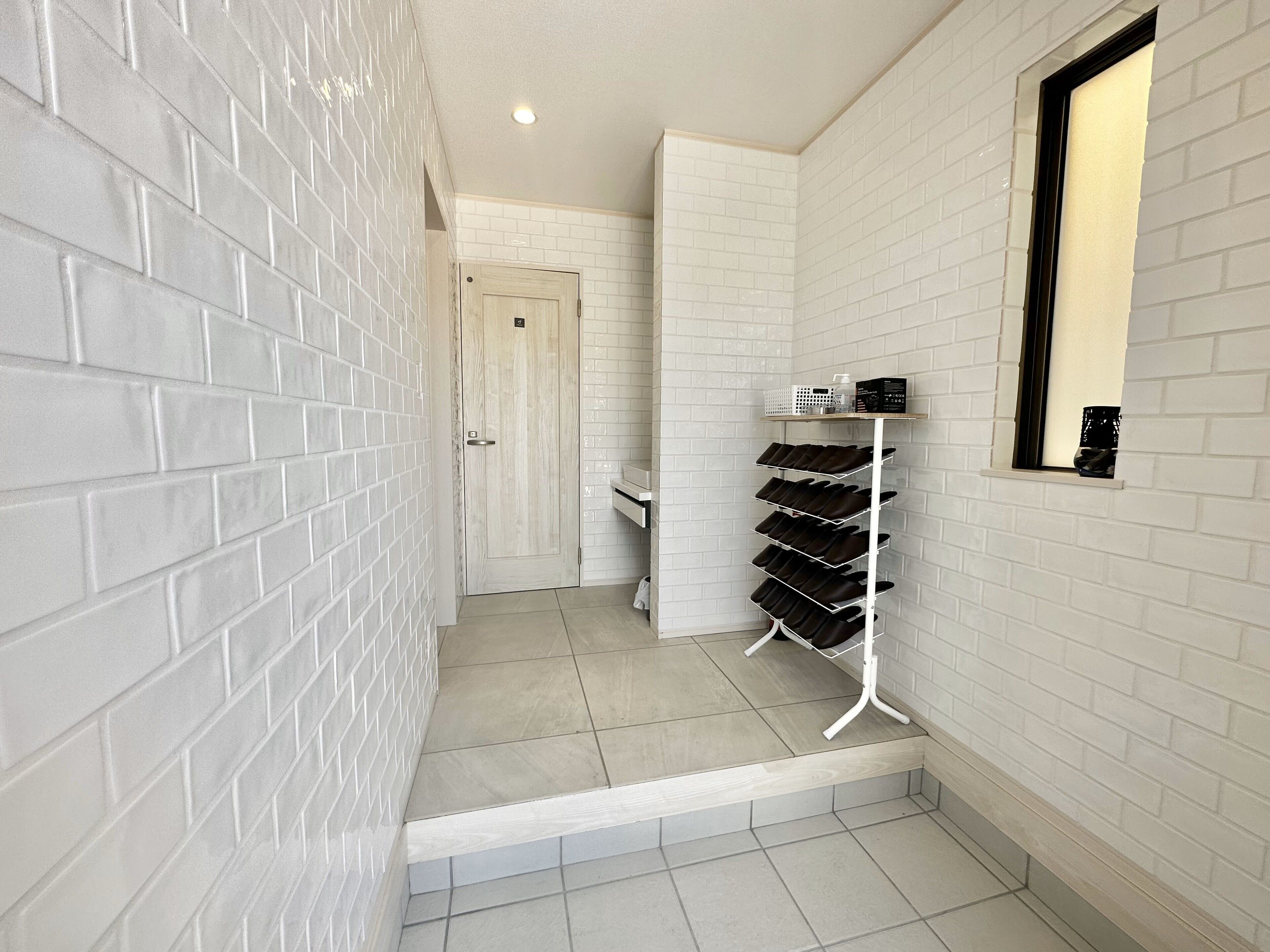 A narrow hallway with white brick walls and a white tiled floor. A white metal rack with shoes is mounted on the wall, and a small window with a black frame is visible.