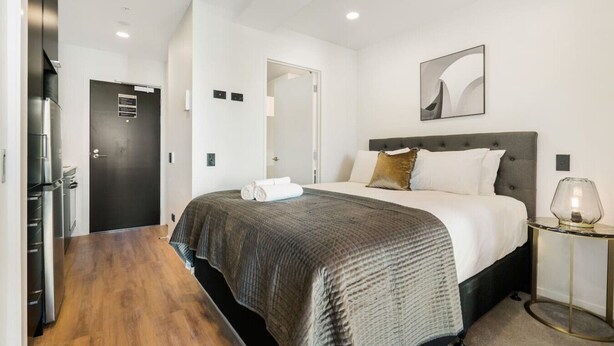 Una habitación de hotel moderna con una cama, una mesita de noche, una lámpara y una puerta que da a una cocina.
