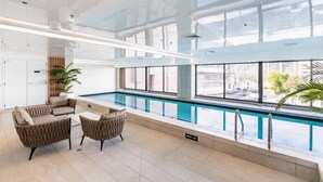 Una piscina interior moderna con paredes de vidrio transparente, un área de descanso con asientos acolchados y vistas a la ciudad.
