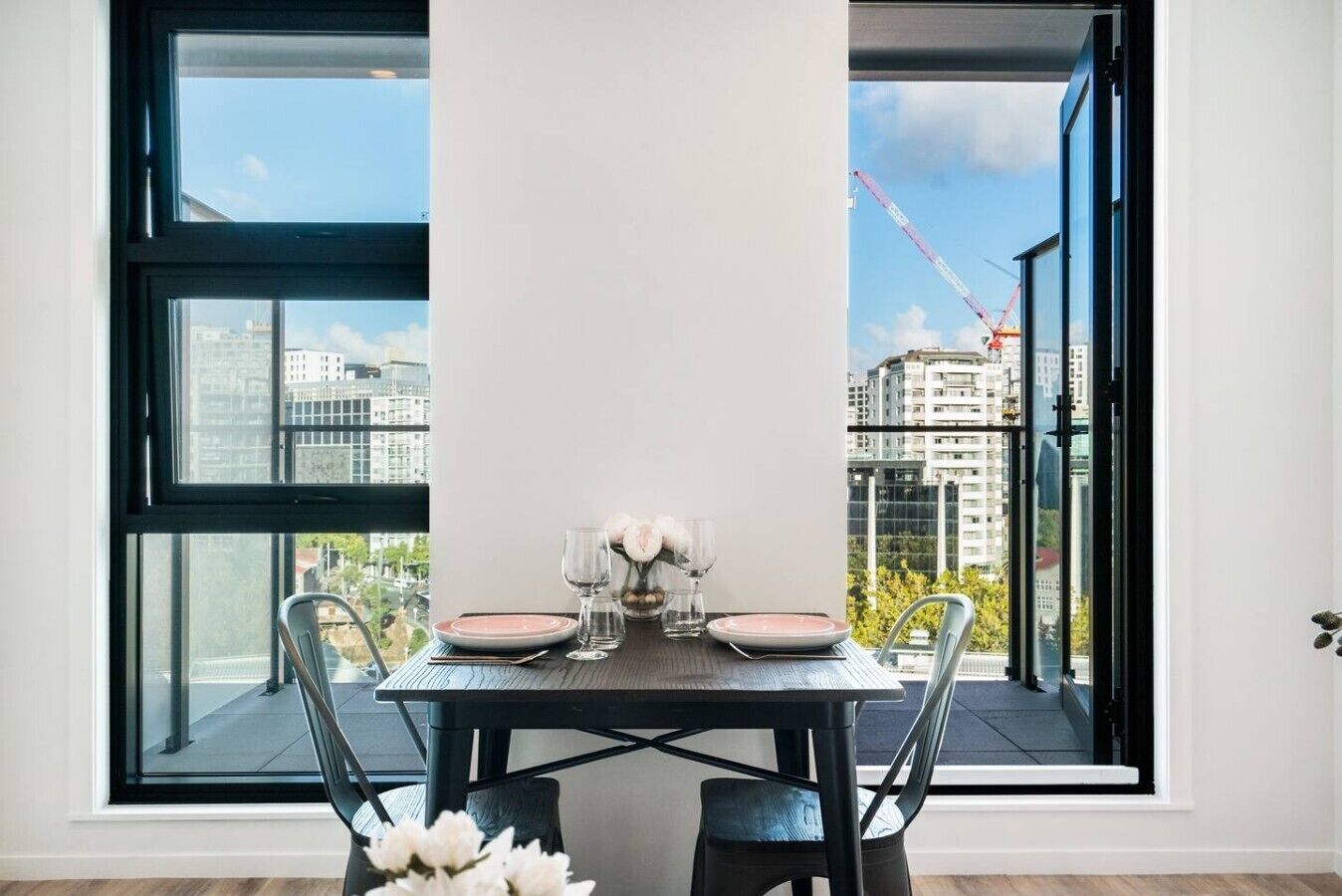 Un coin repas avec une table dressée pour deux personnes, de grandes fenêtres et une vue sur des immeubles et une grue.