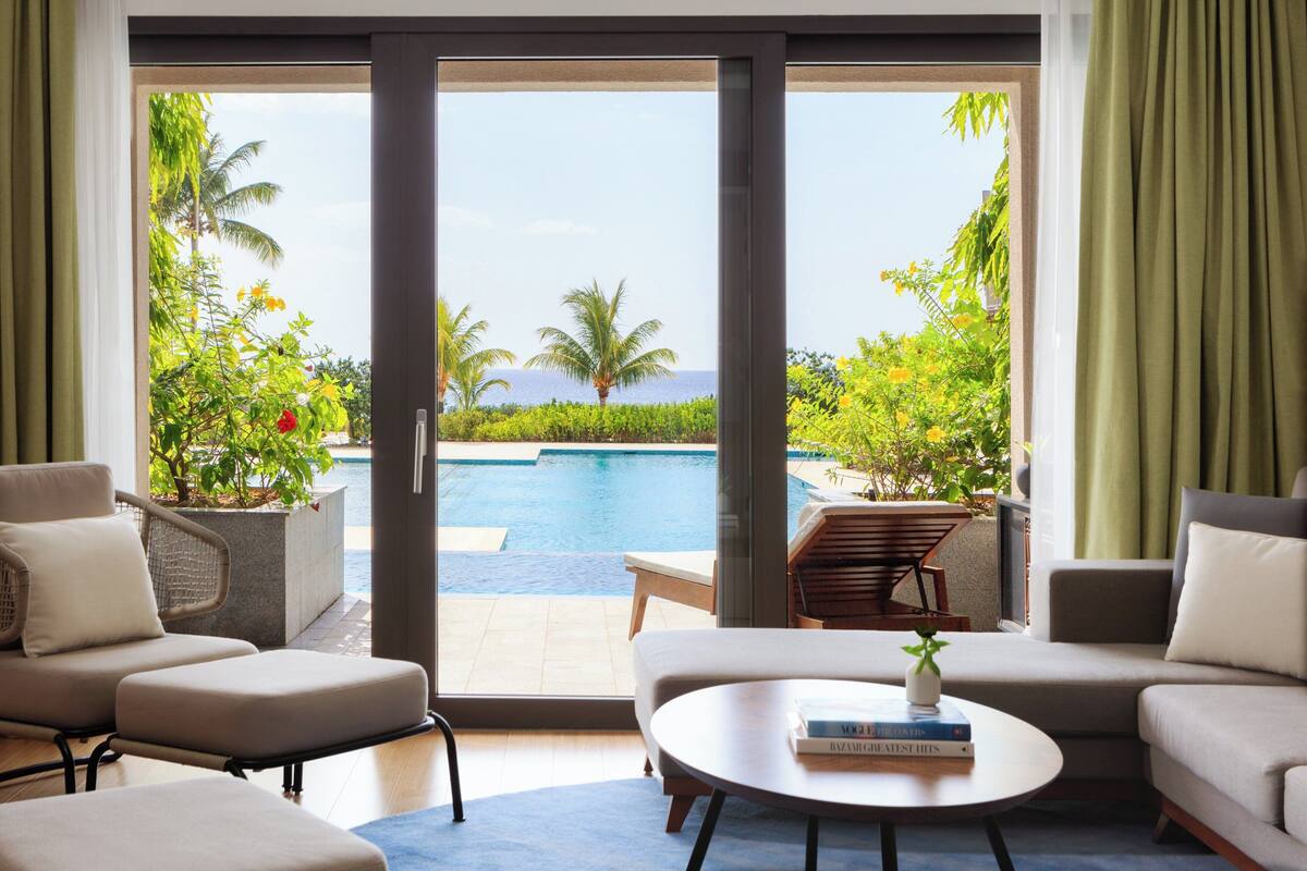 A modern living room with a large sliding glass door leading to a pool area, featuring a sofa, armchairs, and a coffee table.