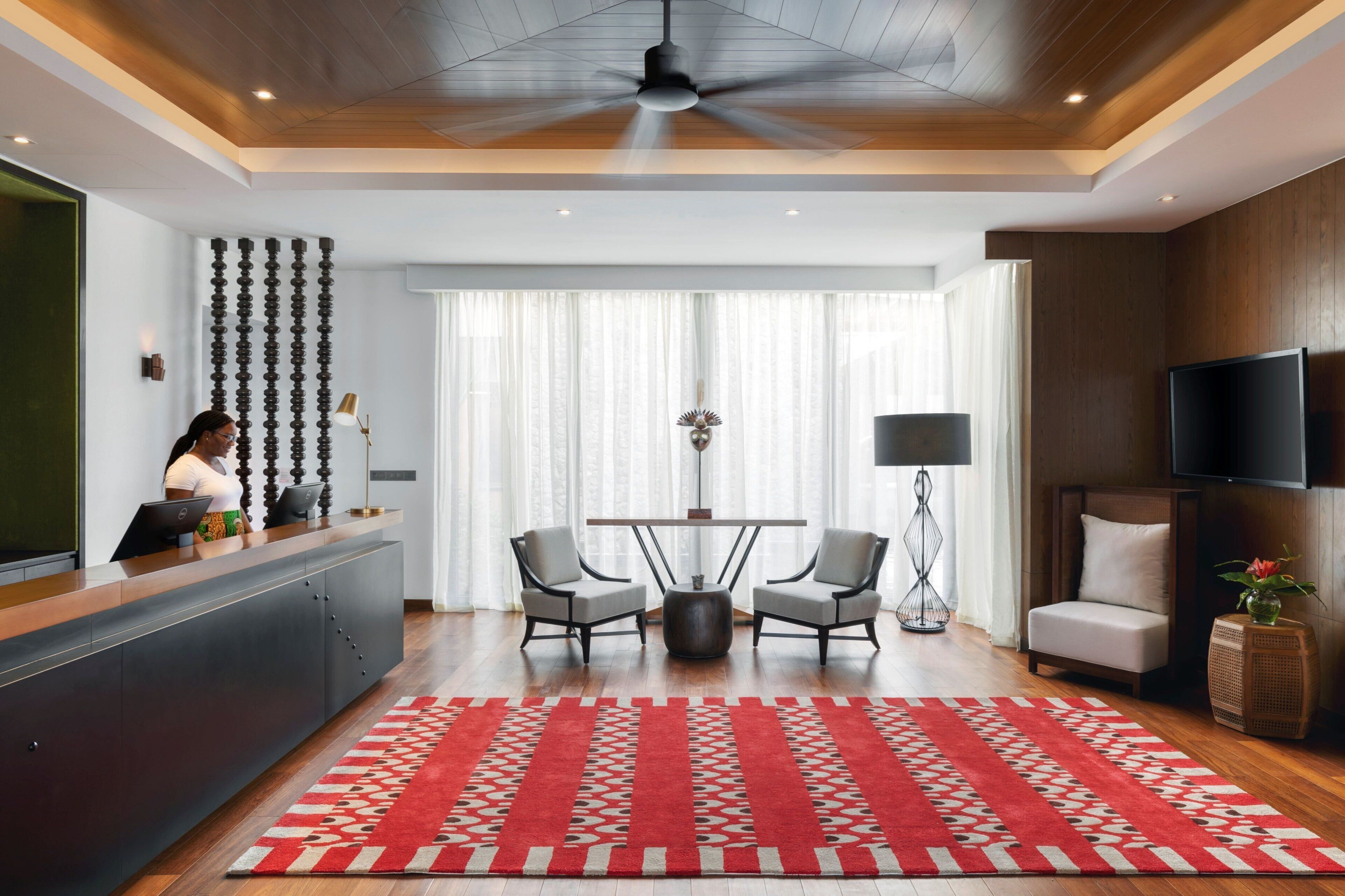 A modern hotel lobby with a reception desk, a seating area with two chairs, a large red patterned rug, and a ceiling fan.