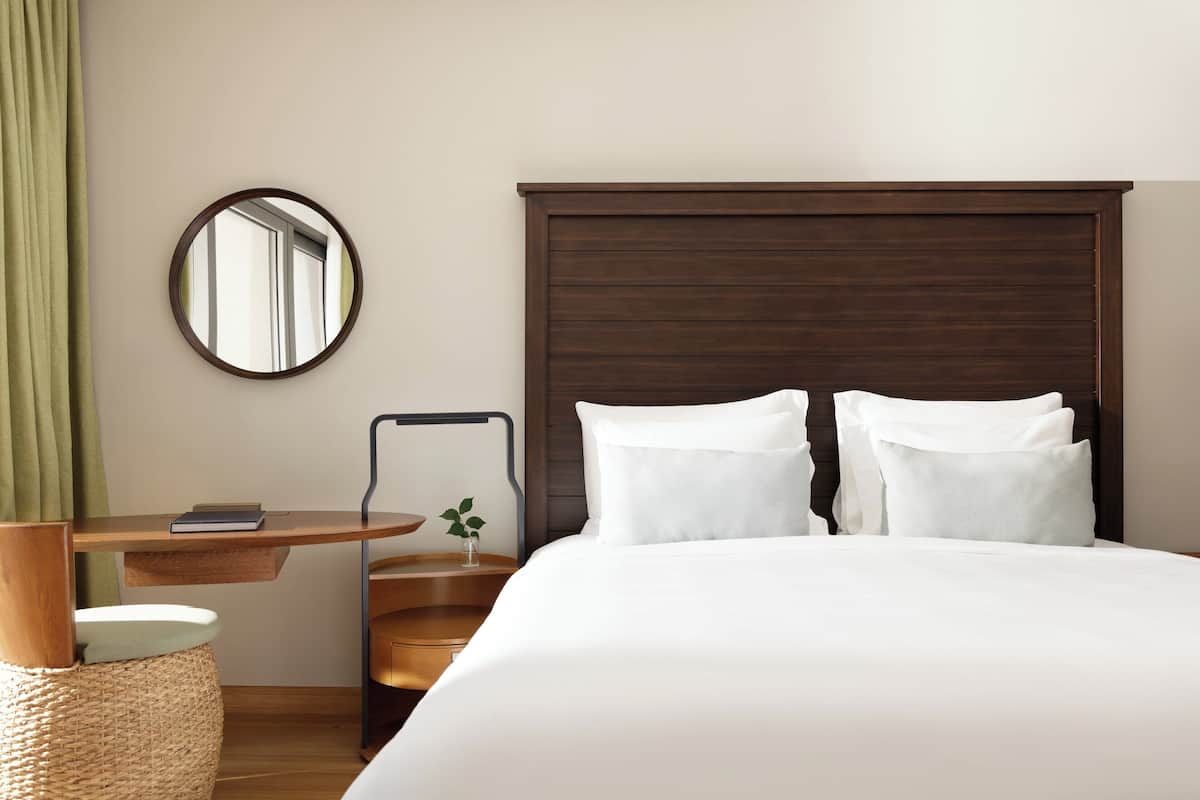 A hotel room with a large bed, a wooden headboard, a round mirror, a small table with a book, and a green curtain.