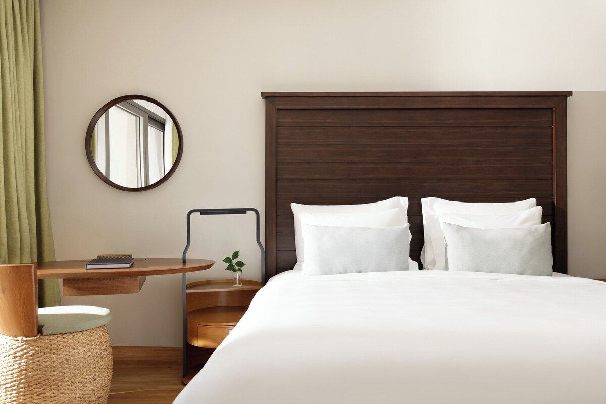 Habitación de hotel con una cama grande, cabecera de madera, espejo redondo, una pequeña mesa con un libro y una cortina verde.