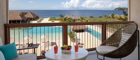 A balcony with a view of a beach and pool, wicker chairs, and a table with drinks and fruit.